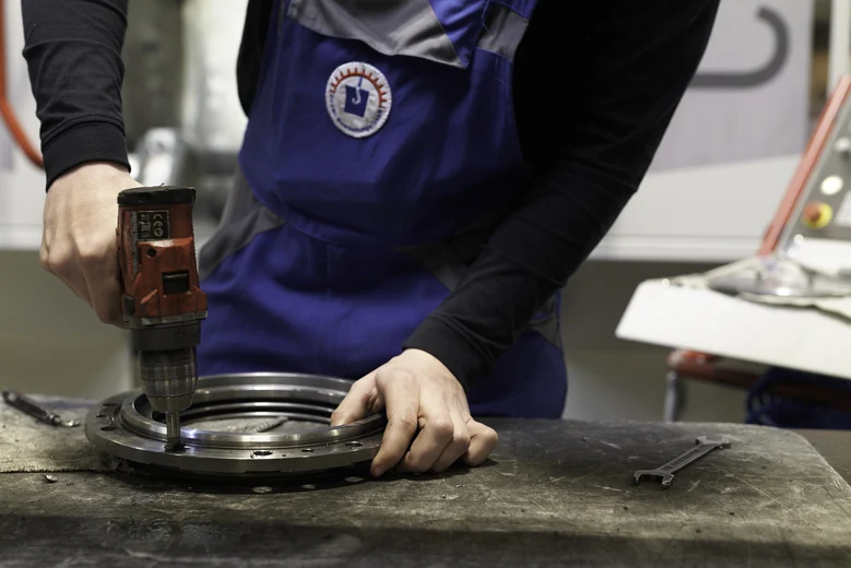 Hände eines Arbeiters an einem Metallwerkstück und einer Bohrmaschine.