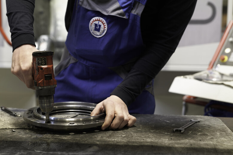Hände eines Arbeiters an einem Metallwerkstück und einer Bohrmaschine.